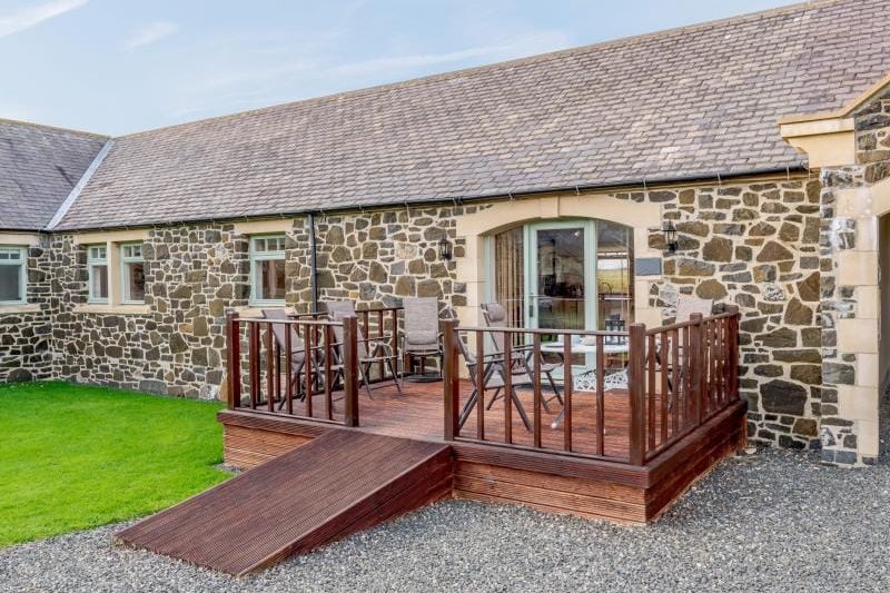 Stone-built cottage with a wooden deck and ramp to a glass door, outdoor seating on the deck.