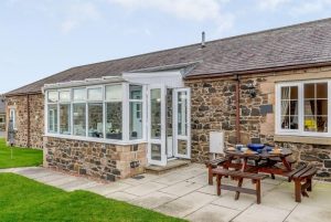 Stone cottage with a white-framed glass conservatory and a wooden picnic table on a paved patio.