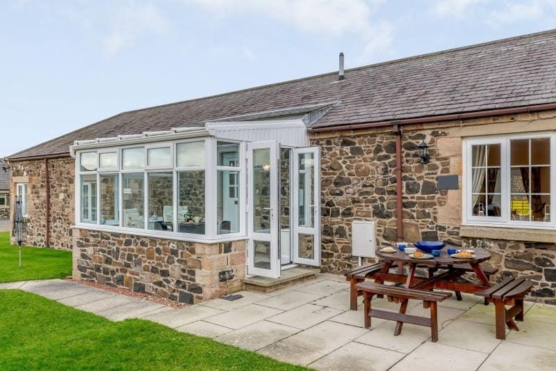 Stone cottage with a white-framed glass conservatory and a wooden picnic table on a paved patio.