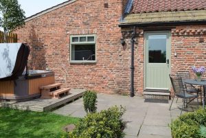 Brick backyard with a hot tub on a wooden deck, steps leading up, and a pale green door beside a window.